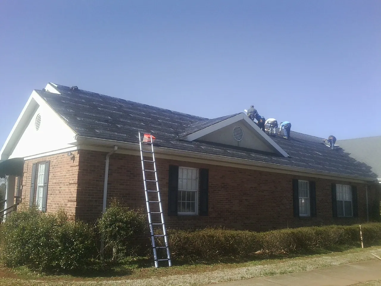 Crew tearing shingles off a brick ranch roof with a ladder set — Metal Roof Repair work by our crew in Ridge