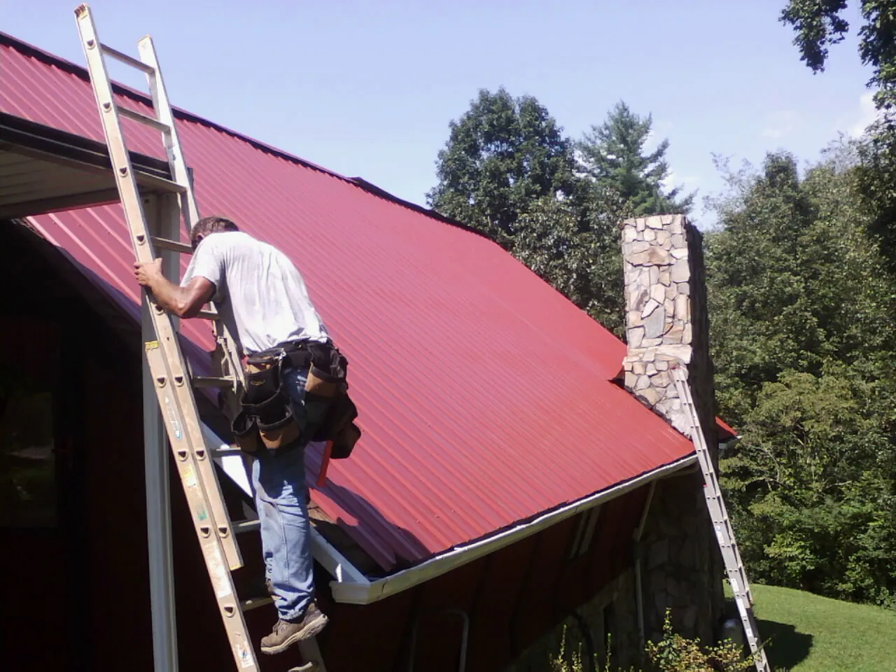 Roofer on a ladder beside a red metal roof and stone chimney — Soffit & Fascia Repair by Roofing Brothers in Ridge
